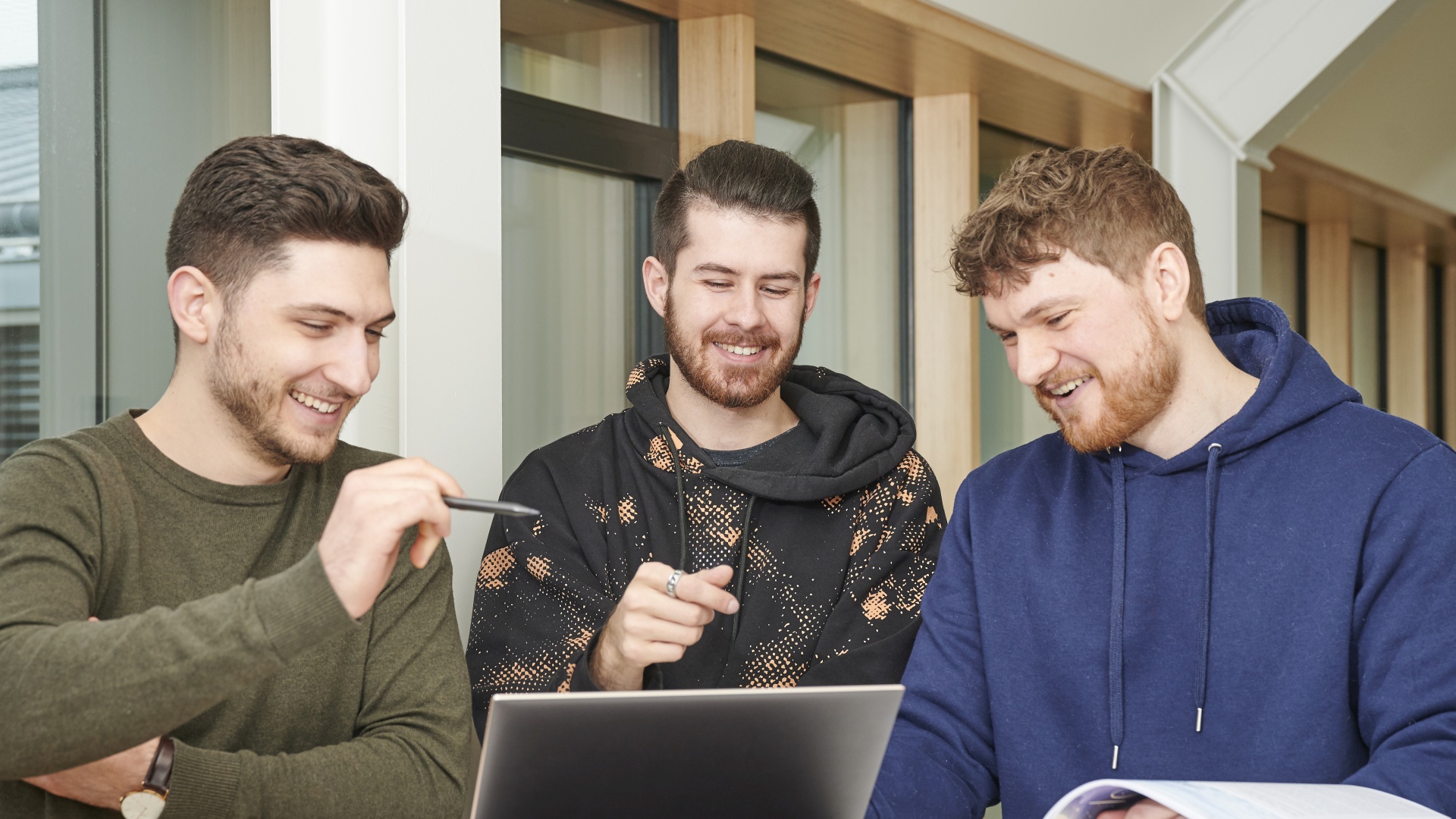 Drei Männer blicken lachend auf einen Laptop. 