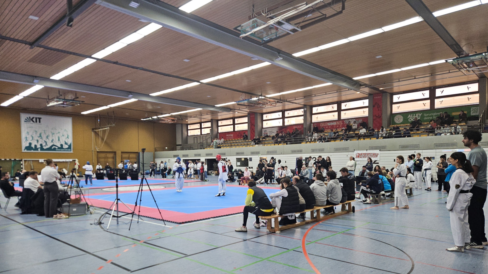 Taekwondo tournament in a gym with competitors, judges, and spectators on mats.