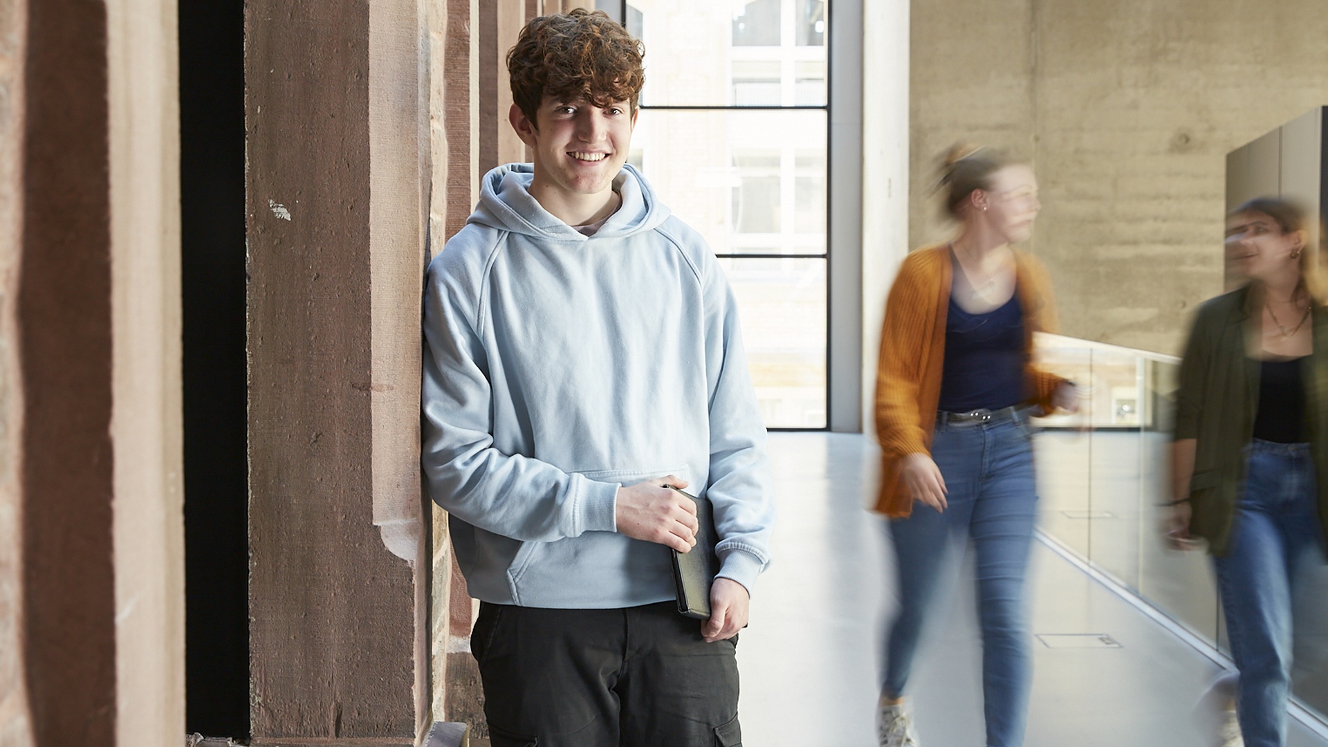 Ein junger Student lehnt lächelnd mit Laptop in der Hand, an eine Wand. 