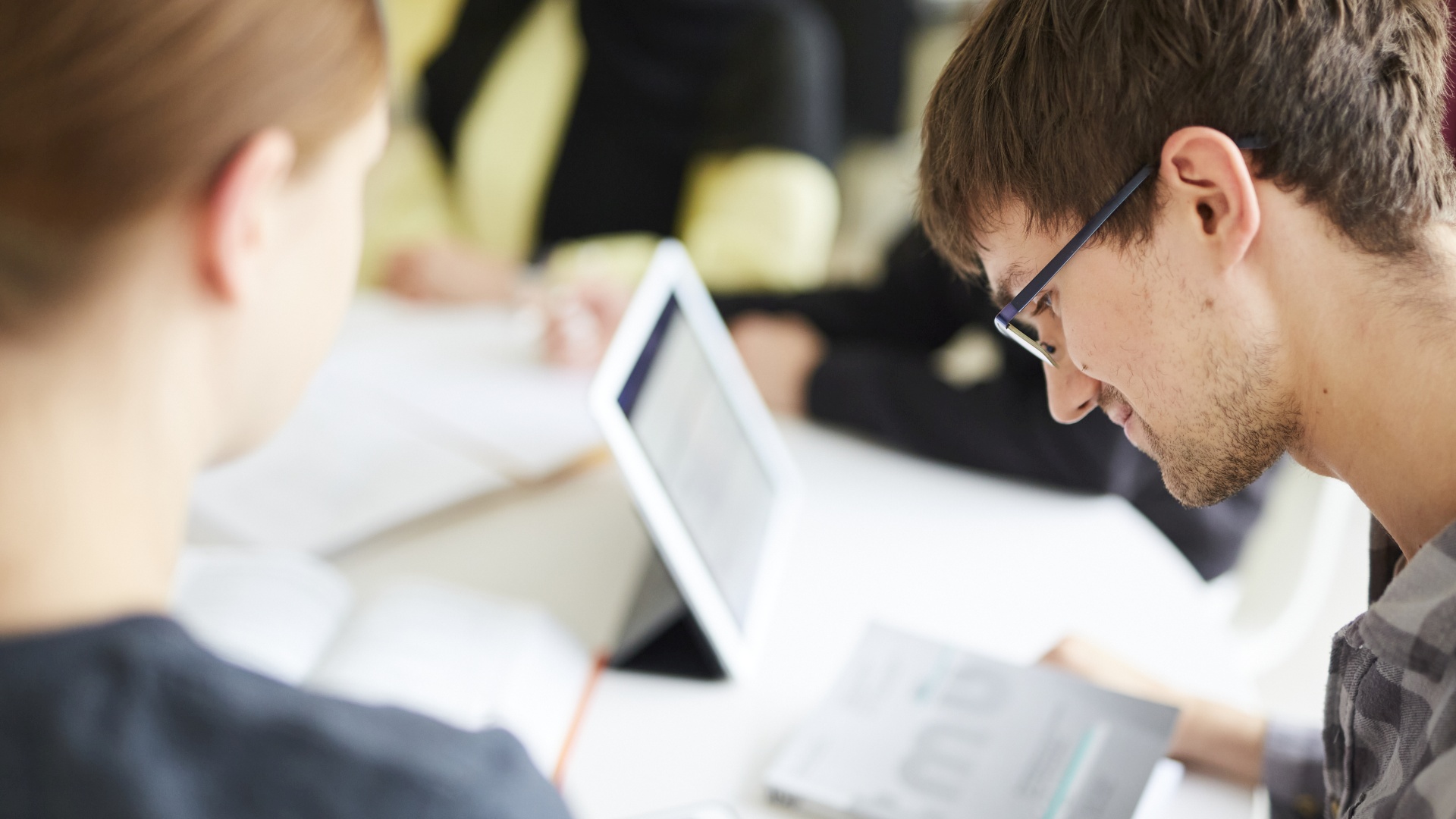 Personen arbeiten gemeinsam mit Tablet und Zeitschrift an einem Tisch in hellem Raum.