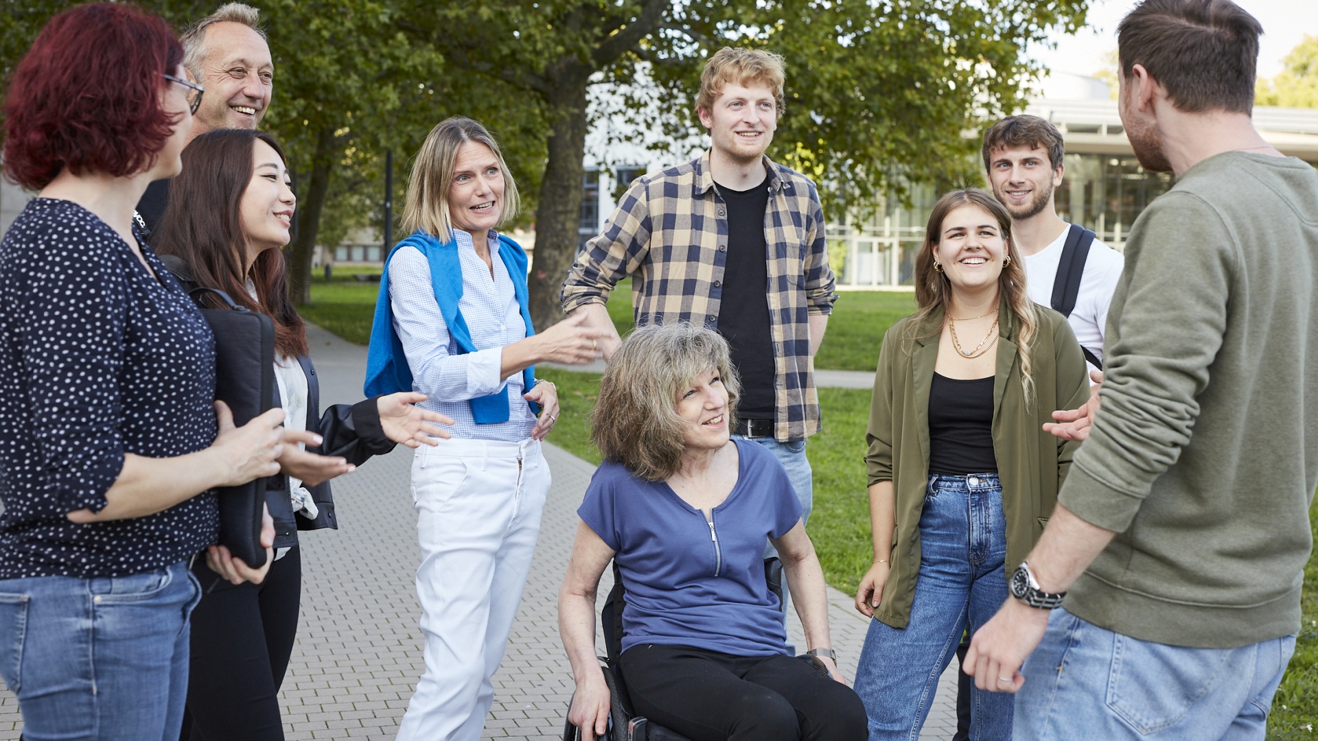 Gruppe junger Menschen um eine sitzende ältere Frau in Rollstuhl, Outdoor auf Campus.