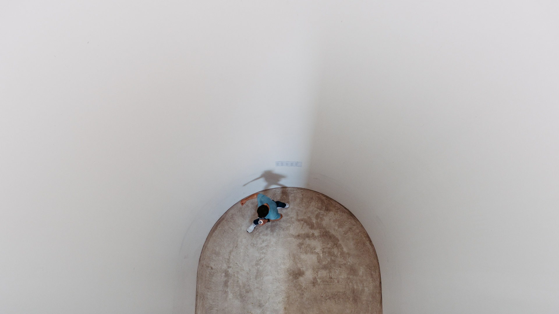 Mann in blauem T-Shirt geht allein in rundem, weißem Raum mit Schatten an der Wand.
