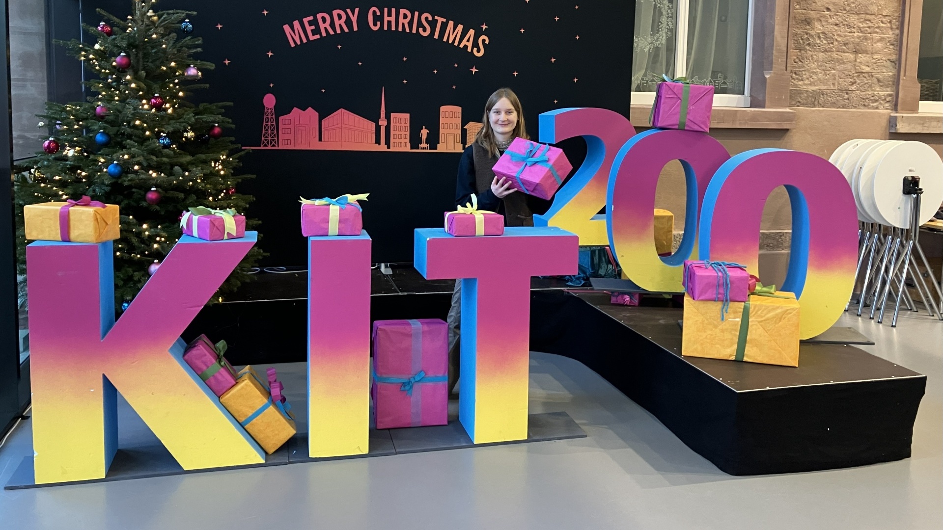 Marina Schreidl steht hinter pink-gelbem KIT-Banner mit einem Geschenk in der Hand.