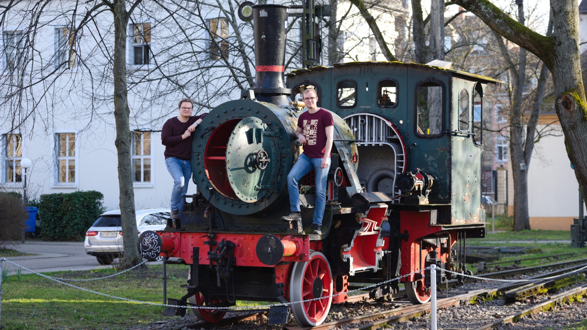 Old steam locomotive on tracks with two people standing on it in a park setting.
