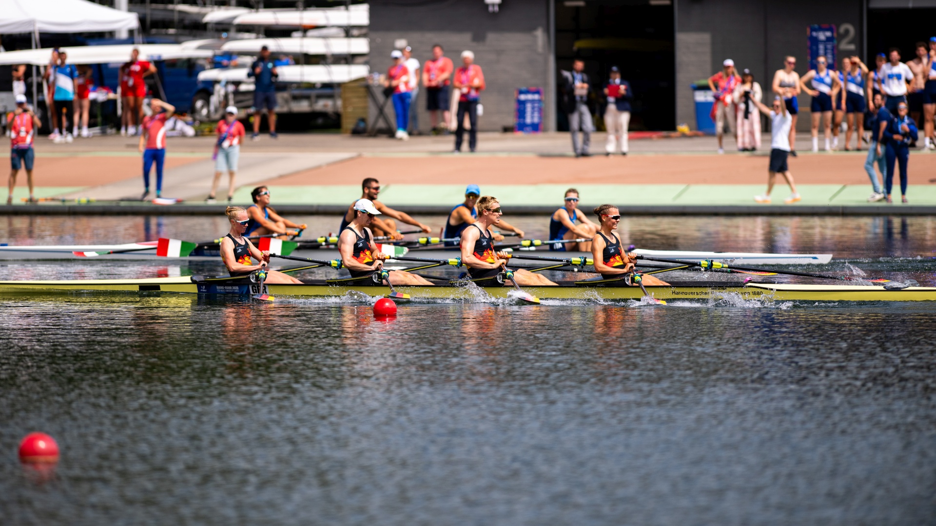 Ruderwettbewerb mit mehreren Mannschaften auf dem Wasser und Zuschauern am Ufer.